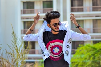 A man with sunglasses is adjusting his white jacket with one hand raised. He is wearing a black t-shirt with a bold circular design featuring red, white, and blue colors. Behind him, there is an outdoor setting with a building and some greenery visible.