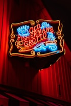 A brightly lit neon sign advertises 'Old Fisherman's Grotto', offering seafood, steaks, and pasta. The sign has vibrant red, blue, and yellow colors that stand out against a dark wooden background.