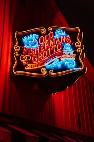 A brightly lit neon sign advertises 'Old Fisherman's Grotto', offering seafood, steaks, and pasta. The sign has vibrant red, blue, and yellow colors that stand out against a dark wooden background.