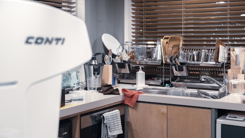 A modern kitchen features a countertop with various kitchen utensils and equipment. The space includes a coffee machine, dish rack filled with plates, glasses, and cutting boards, and a sink area with a soap dispenser. Wooden blinds cover the window in the background, adding warmth to the setting.