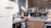 A modern kitchen features a countertop with various kitchen utensils and equipment. The space includes a coffee machine, dish rack filled with plates, glasses, and cutting boards, and a sink area with a soap dispenser. Wooden blinds cover the window in the background, adding warmth to the setting.