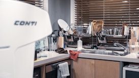 A modern kitchen features a countertop with various kitchen utensils and equipment. The space includes a coffee machine, dish rack filled with plates, glasses, and cutting boards, and a sink area with a soap dispenser. Wooden blinds cover the window in the background, adding warmth to the setting.