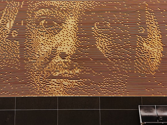 An intricate wall art depicting a face, created using alternating horizontal lines and small squares. The artwork features shades of brown and beige, resulting in a detailed and textured portrait. Below the artwork, there is a tiled floor with a single black bench positioned against the wall.