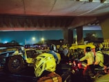 Traffic congestion under an overpass during the evening with vehicles such as cars, motorcycles, and auto-rickshaws. The scene is dimly lit with streetlights and includes several people wearing helmets.