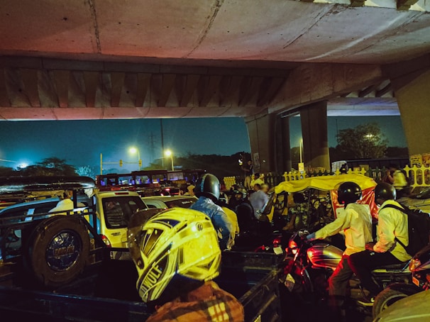 Traffic congestion under an overpass during the evening with vehicles such as cars, motorcycles, and auto-rickshaws. The scene is dimly lit with streetlights and includes several people wearing helmets.