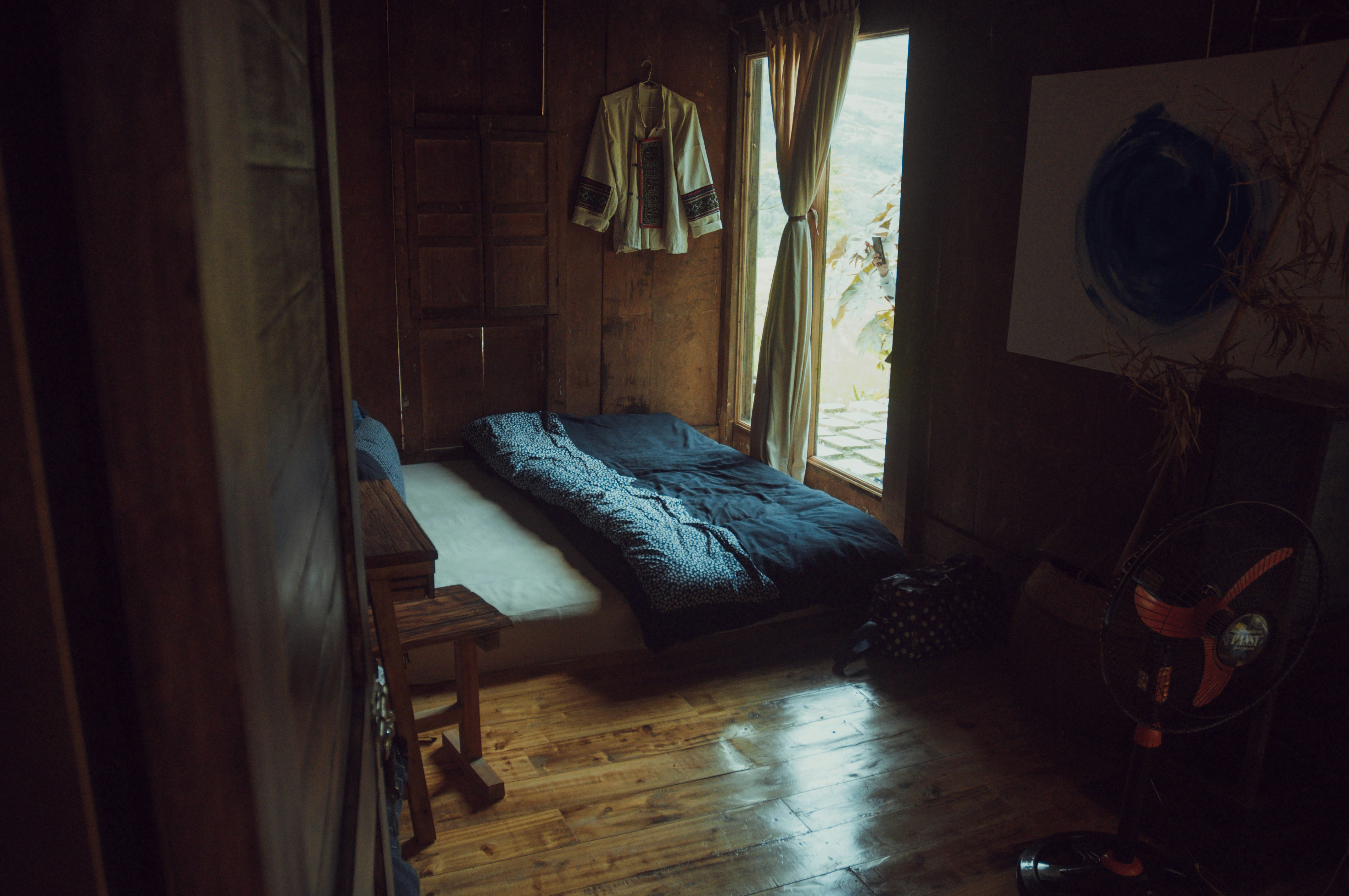 A cozy, rustic room with wooden walls and floor. A single bed is laid out with a dark, patterned duvet. A traditional garment hangs on the wall, and soft light filters through a curtain beside a small window. A standing fan is visible in the corner, next to a wooden table and a decorative piece with bamboo stems.