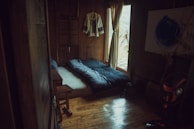 A cozy, rustic room with wooden walls and floor. A single bed is laid out with a dark, patterned duvet. A traditional garment hangs on the wall, and soft light filters through a curtain beside a small window. A standing fan is visible in the corner, next to a wooden table and a decorative piece with bamboo stems.