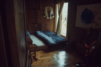 A cozy, rustic room with wooden walls and floor. A single bed is laid out with a dark, patterned duvet. A traditional garment hangs on the wall, and soft light filters through a curtain beside a small window. A standing fan is visible in the corner, next to a wooden table and a decorative piece with bamboo stems.