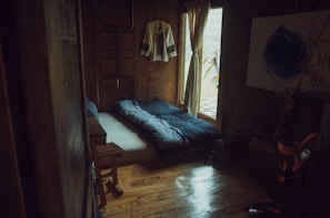 A cozy, rustic room with wooden walls and floor. A single bed is laid out with a dark, patterned duvet. A traditional garment hangs on the wall, and soft light filters through a curtain beside a small window. A standing fan is visible in the corner, next to a wooden table and a decorative piece with bamboo stems.