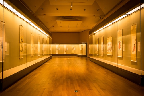 A well-lit gallery room with wooden flooring and glass display cases on both sides containing hanging scrolls. The lighting is warm, creating a serene and contemplative atmosphere. The scrolls feature various traditional artworks and calligraphy.