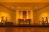 A well-lit museum gallery featuring traditional Asian furniture and artwork. Three vertical scrolls with Asian calligraphy hang on the wall above a central wooden table with decorative elements. Symmetrically placed chairs flank the table, creating a balanced and harmonious setting.
