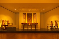 A well-lit museum gallery featuring traditional Asian furniture and artwork. Three vertical scrolls with Asian calligraphy hang on the wall above a central wooden table with decorative elements. Symmetrically placed chairs flank the table, creating a balanced and harmonious setting.