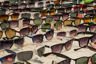 A collection of various sunglasses laid out for display, with different styles and colors prominently featured. Some frames are black, white, blue, and brown, with lenses ranging from dark to light tints. Brand tags are visible on many of the sunglasses.