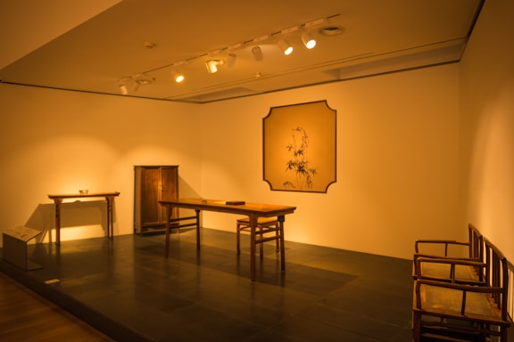 A minimalistic interior scene features traditional Asian furniture within a softly lit room. The setup includes a wooden table, chairs, and a small cabinet against a neutral-colored wall. A framed artwork with bamboo motifs hangs on the wall, enhancing the room's cultural aesthetic. Subtle lighting adds warmth and highlights the simplicity and elegance of the furnishings.