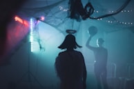 A dimly lit room features a person in a costume with a halo, suggestive of an angel theme, silhouetted against a backdrop of eerie, decorative lighting and faux cobwebs. Another figure holds a ball in a lively, atmospheric scene.