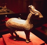 A detailed bronze artifact shaped like an animal, possibly a ceremonial vessel or sculpture, with intricate patterns and designs covering its surface. The piece is displayed on a red fabric platform, suggesting a museum or exhibition setting.