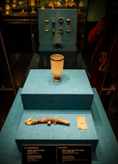 A display case features ancient artifacts arranged on three blue-tiered platforms. The closest platform holds a jade-colored artifact resembling a bracelet and a square-shaped object with intricate designs. The middle platform displays a white cup with gold accents. The background showcases additional artifacts, including rings, in a museum setting.