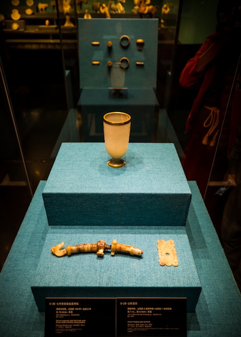 A display case features ancient artifacts arranged on three blue-tiered platforms. The closest platform holds a jade-colored artifact resembling a bracelet and a square-shaped object with intricate designs. The middle platform displays a white cup with gold accents. The background showcases additional artifacts, including rings, in a museum setting.