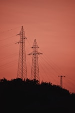 Silhouetted power transmission towers rise against a warm, reddish-orange sky during sunset. The towers are surrounded by minimal foliage at their base, and several power lines stretch between them, adding a geometric element to the scene.