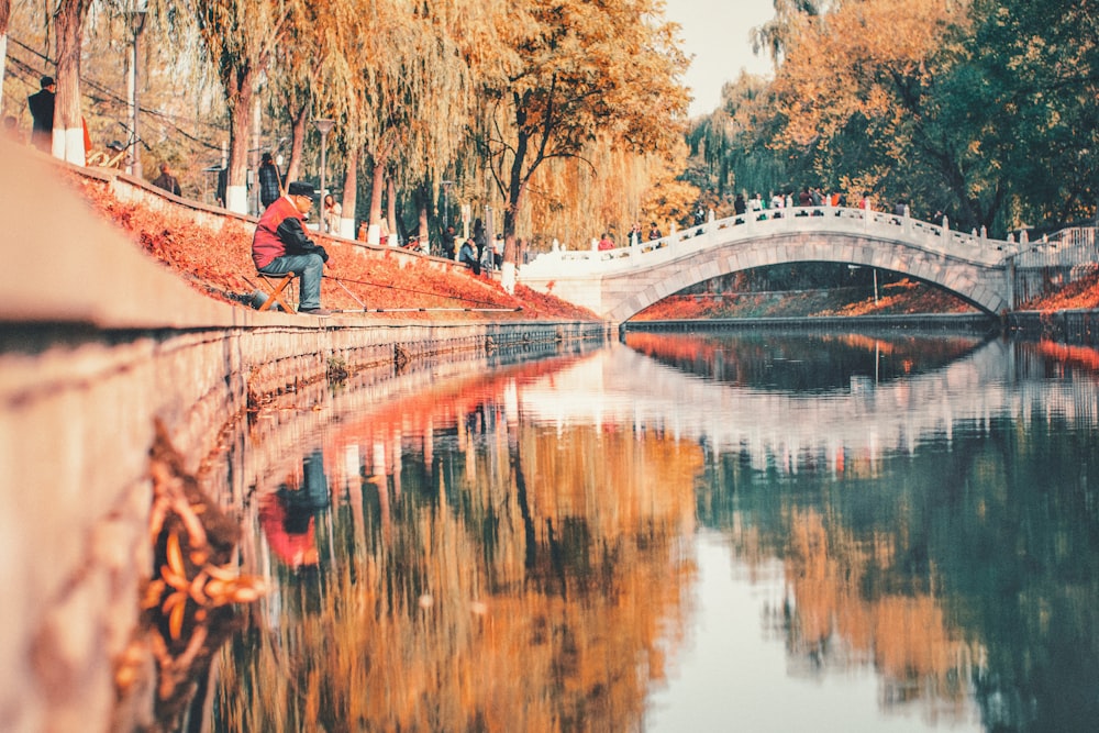 photo of man wearing orange and black jacket sitting on chair beside on river