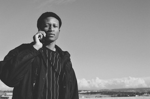 A person is talking on a mobile phone outdoors. They are wearing a dark jacket over a striped shirt, with a bright sky and distant landscape in the background. The image is in black and white.