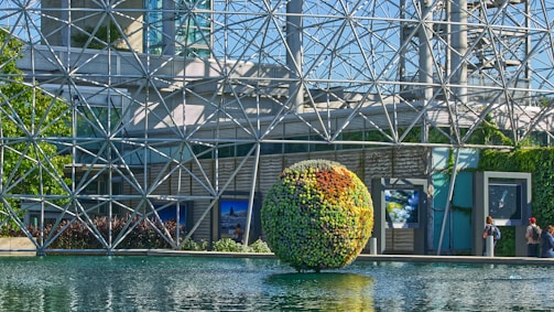 A large geodesic dome made of metallic struts forms the backdrop of a spherical structure covered in greenery, floating on a serene body of water. The structure reflects the sunlight creating a vivid display of colors. In the foreground, a few people are observing the scene, adding a sense of scale and human presence.