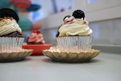 Cupcakes with decorative frosting are presented on small golden plates. The main cupcake features white whipped cream frosting with a blackberry on top. Other cupcakes in the background display different frosting colors like pink and brown.