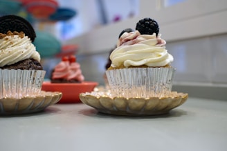 Cupcakes with decorative frosting are presented on small golden plates. The main cupcake features white whipped cream frosting with a blackberry on top. Other cupcakes in the background display different frosting colors like pink and brown.