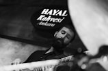 A black and white image featuring a man playing a bass guitar. The man appears focused and engaged with the instrument. In the background, a sign reads 'Hayal Kahvesi Ankara'. The lighting and shadows create a moody and artistic atmosphere.