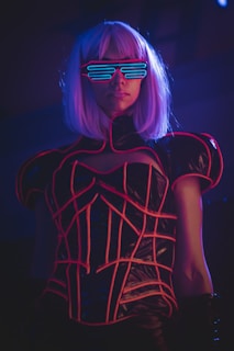 A person with a short white wig is wearing a futuristic outfit featuring neon pink and blue elements. The person is adorned with light-up glasses that have horizontal blue lines across the lenses, creating a cyberpunk aesthetic.