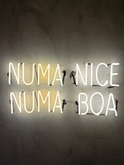 Neon lights display the words NUMA and NICE on the top row and NUMA and BOA on the bottom row against a textured dark wall.