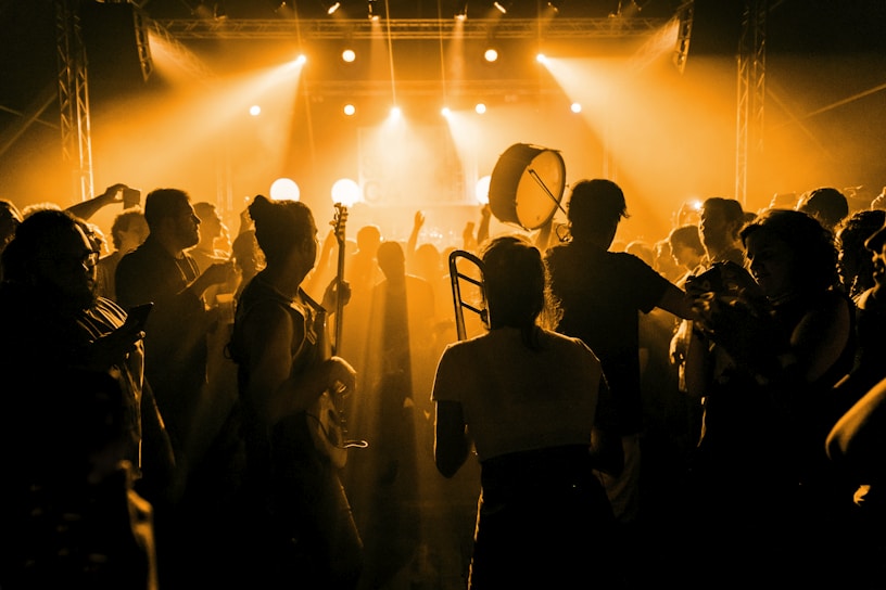 A crowded concert scene with silhouettes of people enjoying live music. The dimly lit setting is enhanced by warm yellow lighting that creates a vibrant atmosphere. Some individuals carry musical instruments like a trombone and a drum, indicating live performance involvement.