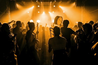 A crowded concert scene with silhouettes of people enjoying live music. The dimly lit setting is enhanced by warm yellow lighting that creates a vibrant atmosphere. Some individuals carry musical instruments like a trombone and a drum, indicating live performance involvement.