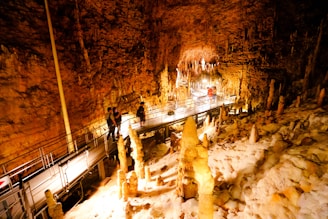 A mesmerizing cave interior with striking stalagmites and stalactites illuminated by warm lighting. A metal walkway with railings accommodates several people as they explore the natural formations.