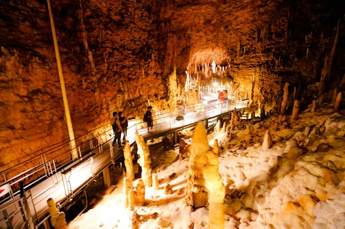 A mesmerizing cave interior with striking stalagmites and stalactites illuminated by warm lighting. A metal walkway with railings accommodates several people as they explore the natural formations.