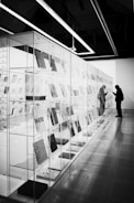In an exhibition space, a glass-enclosed display of books or documents stretches along the wall. A person, silhouetted and wearing a hat and a backpack, stands closely observing the contents inside the glass case. The room's lighting is bright with overhead fluorescent lights, creating strong contrasts and reflections.