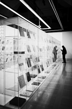 In an exhibition space, a glass-enclosed display of books or documents stretches along the wall. A person, silhouetted and wearing a hat and a backpack, stands closely observing the contents inside the glass case. The room's lighting is bright with overhead fluorescent lights, creating strong contrasts and reflections.