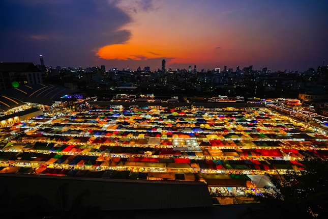 Colorful market stalls are illuminated below a vibrant and dramatic sunset, with tall city buildings silhouetted against the evening sky.