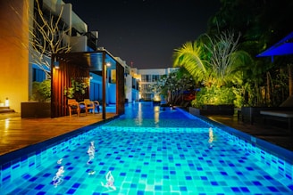 A modern outdoor swimming pool scene at night with vibrant blue lighting reflecting on the water. The pool area is surrounded by lush greenery and contemporary seating areas. Warm lighting adds a cozy ambiance to the setting, and tall palm trees are visible to the right.