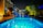 A modern outdoor swimming pool scene at night with vibrant blue lighting reflecting on the water. The pool area is surrounded by lush greenery and contemporary seating areas. Warm lighting adds a cozy ambiance to the setting, and tall palm trees are visible to the right.