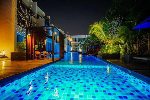 A modern outdoor swimming pool scene at night with vibrant blue lighting reflecting on the water. The pool area is surrounded by lush greenery and contemporary seating areas. Warm lighting adds a cozy ambiance to the setting, and tall palm trees are visible to the right.