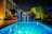 A modern outdoor swimming pool scene at night with vibrant blue lighting reflecting on the water. The pool area is surrounded by lush greenery and contemporary seating areas. Warm lighting adds a cozy ambiance to the setting, and tall palm trees are visible to the right.