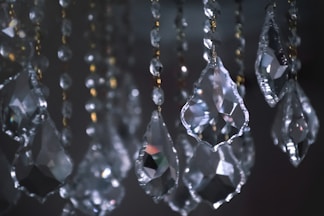A collection of hanging crystal prisms with faceted surfaces, reflecting light and creating small sparkles. The prisms are attached to strands of smaller beads, and there are hints of metallic elements in the design.