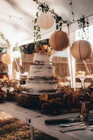 A rustic, three-tiered cake is elegantly decorated with autumn-themed foliage and berries. It sits on a wooden table under a canopy, surrounded by hanging lanterns and green vines. The setting appears to be an outdoor event, possibly a wedding or celebration, with a warm and cozy ambiance.