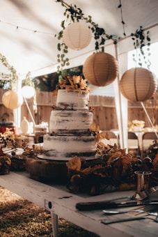 A rustic, three-tiered cake is elegantly decorated with autumn-themed foliage and berries. It sits on a wooden table under a canopy, surrounded by hanging lanterns and green vines. The setting appears to be an outdoor event, possibly a wedding or celebration, with a warm and cozy ambiance.