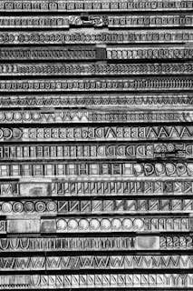 Lines of vintage metal typesetting letters arranged in trays, showcasing a variety of fonts and sizes. The image focuses on the intricate details and textures of the letters, which include uppercase alphabets and symbols in a monochrome style.