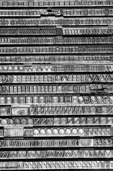 Lines of vintage metal typesetting letters arranged in trays, showcasing a variety of fonts and sizes. The image focuses on the intricate details and textures of the letters, which include uppercase alphabets and symbols in a monochrome style.