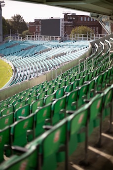 A large outdoor sports stadium with rows of empty green and aqua seats curving around the field. The stadium is well-lit, suggesting it might be daytime. In the background, there is a large digital screen and some buildings, along with trees under a clear sky.