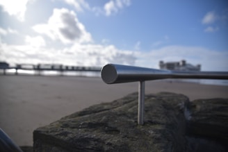 A metal handrail is prominently positioned in the foreground, mounted on a stone structure. The background features a sandy beach leading to a pier extending across the water. The sky is clear with scattered clouds, and the overall atmosphere appears calm and serene.