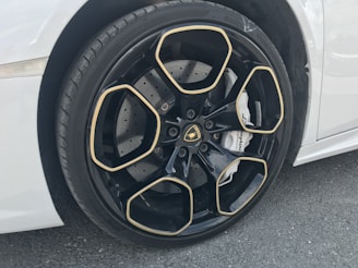 A close-up view of a luxury car wheel featuring a glossy black rim with gold accents in a geometric pattern. The tire has visible tread, and the car is white. The brake caliper displays branding.